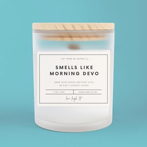 Könnte beinhalten: Ein weißer, mattierter Glas-Kerzenbehälter mit einem Holzdeckel. Das Etikett lautet "Smells Like Morning Devo" und "Grab your coffee and post a pic, or else it doesn't count." Das Etikett enthält auch "11 oz / 120 $", "Vegan, Hergestellt in den USA", und "love laugh lit".