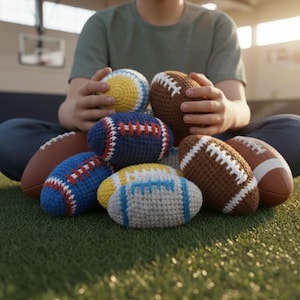 May include: A selection of handmade crochet footballs in various colours, including brown, blue, yellow, and grey. The footballs are arranged on green grass, with one held in the hands of a person. The crochet work features detailed stitching.