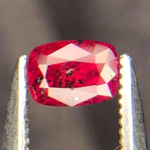 Peut inclure: Une pierre précieuse taille coussin d'une couleur rouge vive avec des inclusions sombres visibles sur toute sa surface à facettes. La pierre est maintenue par une pince en métal argenté aux bords dentelés, mettant en valeur sa clarté.