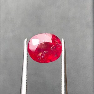 May include: A faceted oval-shaped red gemstone held by silver-toned metal tweezers. The stone displays a vibrant crimson hue with visible internal inclusions and natural surface textures throughout its translucent body, reflecting light on its top facets.