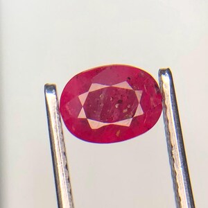 May include: A faceted, oval-shaped ruby gemstone. The ruby is a deep red color with a star-like pattern in the center. The gemstone is held by silver tweezers against a white background.