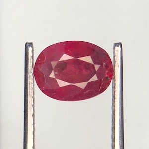 May include: A faceted, oval-shaped ruby gemstone, held by silver tweezers. The ruby is a deep red color with internal facets that create a starburst pattern. The gemstone is set against a white background.