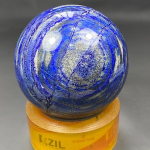 May include: A polished lapis lazuli sphere, showcasing a deep blue hue with streaks of white and gold. The gemstone ball rests on a yellow and orange cylindrical base with the text "LZIL TOKYO".