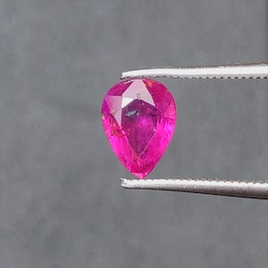 May include: A vibrant, pear-shaped loose gemstone with a deep magenta-pink hue and faceted surface. The stone is held by a pair of silver-toned metal tweezers, showcasing its translucent quality and internal inclusions visible through the facets.
