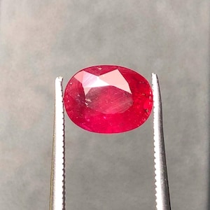 May include: A vibrant, oval-cut red gemstone held securely by a pair of silver-toned metal tweezers. The faceted stone displays a deep, saturated crimson hue with light reflecting off its polished surface, showcasing its clear, transparent structure.