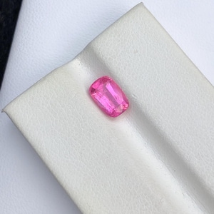 Könnte beinhalten: Ein lebendiger, rechteckig geschliffener rosa Edelstein. Der Edelstein liegt auf einer weißen Oberfläche und zeigt seine leuchtende Farbe und Klarheit. Die Facetten des Steins reflektieren das Licht und erzeugen einen funkelnden Effekt. Ideal für Schmuckherstellung oder Sammlungen.