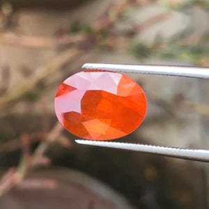 Puede incluir: Una gema facetada de forma ovalada, en tonos naranja y rojo, es sostenida por unas pinzas. La gema tiene una superficie pulida con múltiples facetas, que reflejan la luz. El fondo está borroso, con toques de verde y marrón.