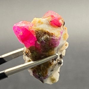 Puede incluir: Una gema de rubí en bruto con vibrantes estructuras cristalinas rosas y rojas. El rubí se encuentra sobre un fondo de minerales blancos, marrones y grises. Sujeta por un par de pinzas plateadas, la gema se muestra sobre un fondo gris neutro.
