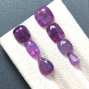 Op de afbeelding: Een verzameling van vijf gefacetteerde paarse edelstenen gerangschikt in twee verticale rijen. De stenen variëren in vorm, waaronder kussen-, ovaal- en peervormige slijpvormen, met diepe magenta en violette tinten en zichtbare interne insluitingen.