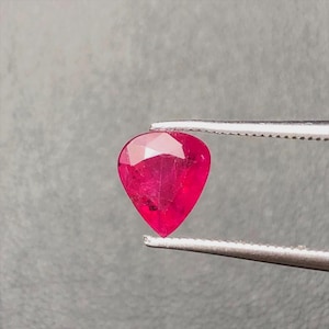 May include: A pear-shaped, faceted gemstone with a vibrant, deep red color. The stone is held by a pair of silver-toned metal tweezers, showcasing its translucent quality and the geometric cut of its surface, which reflects light across its facets.