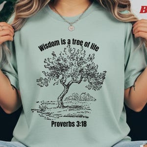 Könnte beinhalten: Salbeigrünes T-Shirt mit einer schwarzen Baumillustration und dem Text "Wisdom is a tree of life" und "Proverbs 3:18". Das Shirt besteht aus einem weichen Material.