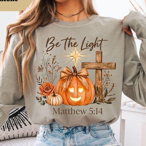 May include: A sandstone-colored long-sleeve sweatshirt featuring a graphic print with a pumpkin, a wooden cross, a star, and floral elements. The text reads Be the Light and Matthew 5:14. The design includes a bow on the pumpkin.