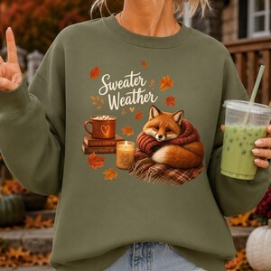 May include: An olive green crewneck sweatshirt featuring a graphic print of a fox wrapped in a scarf, a stack of books, a mug, a candle, and falling leaves. The text Sweater Weather is printed above the fox in a white script font.