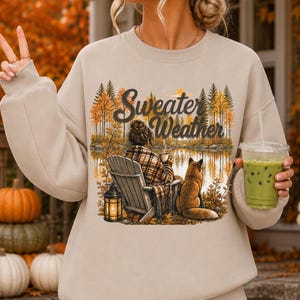 May include: A beige crewneck sweatshirt featuring a graphic print of a person sitting in a wooden chair by a lake with a fox, a lantern, and trees. The text Sweater Weather is printed above the scene. The sweatshirt has long sleeves and a ribbed collar.
