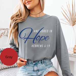 以下が含まれることがあります： 青と白で「Anchored in Hope Hebrews 6:19」の文字が入ったグレーの長袖シャツ。柔らかい素材でできており、クルーネックです。シャツは無地のグレーです。
