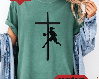 May include: A light green t-shirt featuring a black graphic print of a basketball player in motion positioned in front of a vertical cross. The shirt has short sleeves and a crew neckline. The text "light green" and "comfort colors" appear.