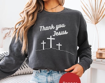 May include: A charcoal gray long-sleeve crewneck sweatshirt featuring white screen-printed text that reads Thank you Jesus above a graphic illustration of three crosses standing on a hill. The garment has a ribbed collar and cuffs.