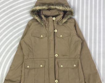 May include: A tan winter jacket with a hood trimmed with faux fur. The jacket has four front pockets, button closures, and a small logo near the bottom. The jacket is designed for cold weather and is made of a durable material.