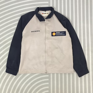 Könnte beinhalten: Beige und marineblaue Jacke mit Reißverschluss und dem Logo "HONDA" auf der Brust. Die Jacke hat einen "Shell SENSE" Aufnäher und ein "RISING SUN" Logo am Kragen. Die Jacke befindet sich auf einem gestreiften Hintergrund.