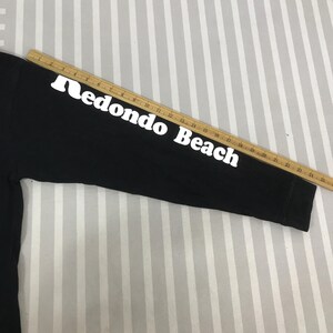 May include: Black long-sleeved shirt with the words "Redondo Beach" printed in white on the sleeve. The shirt is made of a soft material and is suitable for casual wear. A wooden ruler is placed on top of the sleeve.