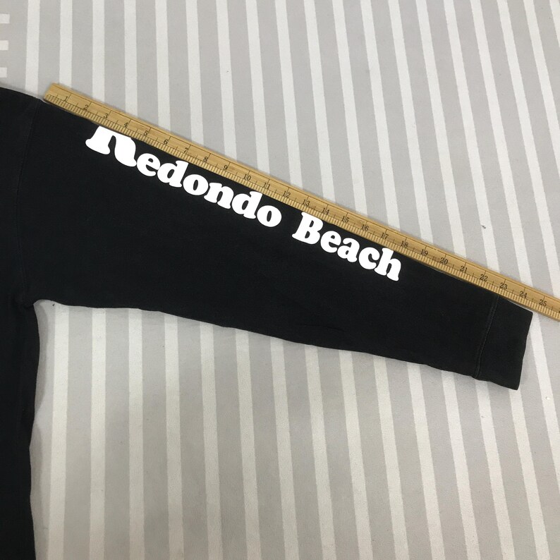 May include: Black long-sleeved shirt with the words "Redondo Beach" printed in white on the sleeve. The shirt is made of a soft material and is suitable for casual wear. A wooden ruler is placed on top of the sleeve.