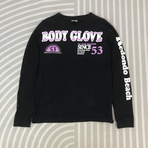 May include: Black long-sleeve shirt with the text "BODY GLOVE" in white and purple. The shirt also features the text "IN THE WATER SINCE 53" and "Redondo Beach" on the sleeve. The shirt has a crew neck.