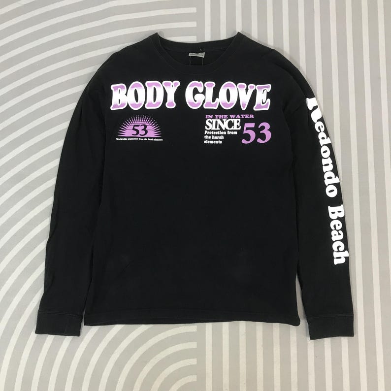 May include: Black long-sleeve shirt with the text "BODY GLOVE" in white and purple. The shirt also features the text "IN THE WATER SINCE 53" and "Redondo Beach" on the sleeve. The shirt has a crew neck.