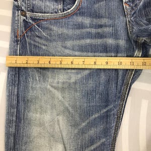 May include: Close-up of blue denim jeans with a wooden ruler measuring the fabric. The jeans have a faded wash and orange stitching. The ruler shows a measurement of approximately 12 inches.