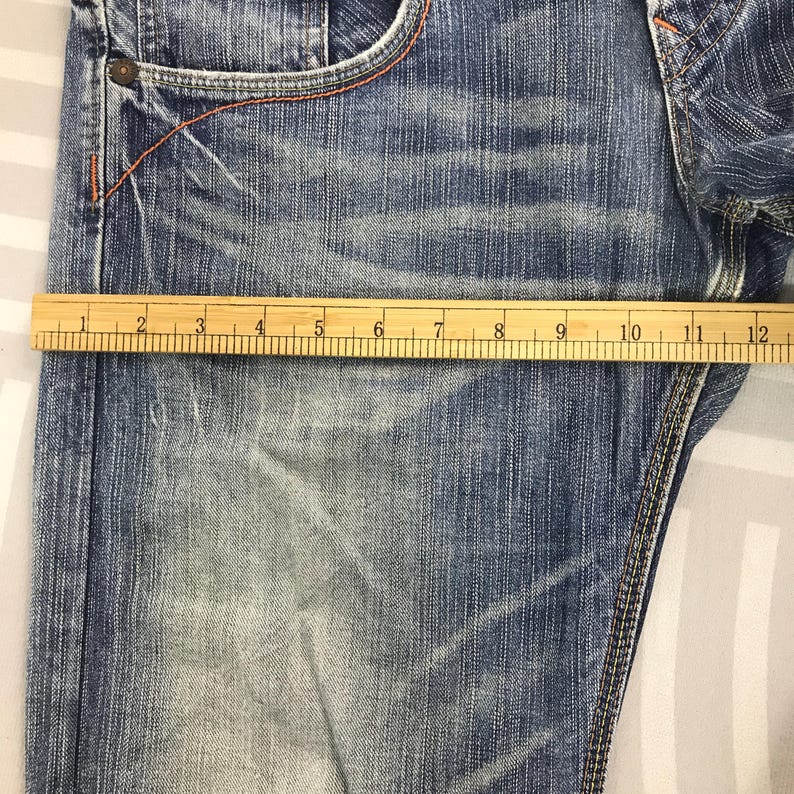 May include: Close-up of blue denim jeans with a wooden ruler measuring the fabric. The jeans have a faded wash and orange stitching. The ruler shows a measurement of approximately 12 inches.
