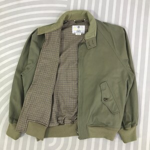 May include: Olive green bomber jacket with a light green collar and cuffs. The jacket has a brown and white checkered lining and a front zipper. A small pocket is visible on the front. The jacket has a label with text.