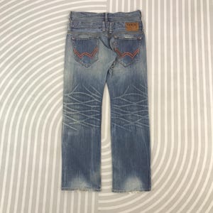 May include: Faded blue denim jeans with red stitching on the back pockets. The jeans have a light wash and a decorative pattern on the legs. A brown leather patch with the brand name "Edwin" is visible.