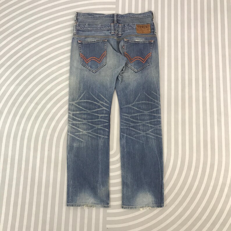 May include: Faded blue denim jeans with red stitching on the back pockets. The jeans have a light wash and a decorative pattern on the legs. A brown leather patch with the brand name "Edwin" is visible.