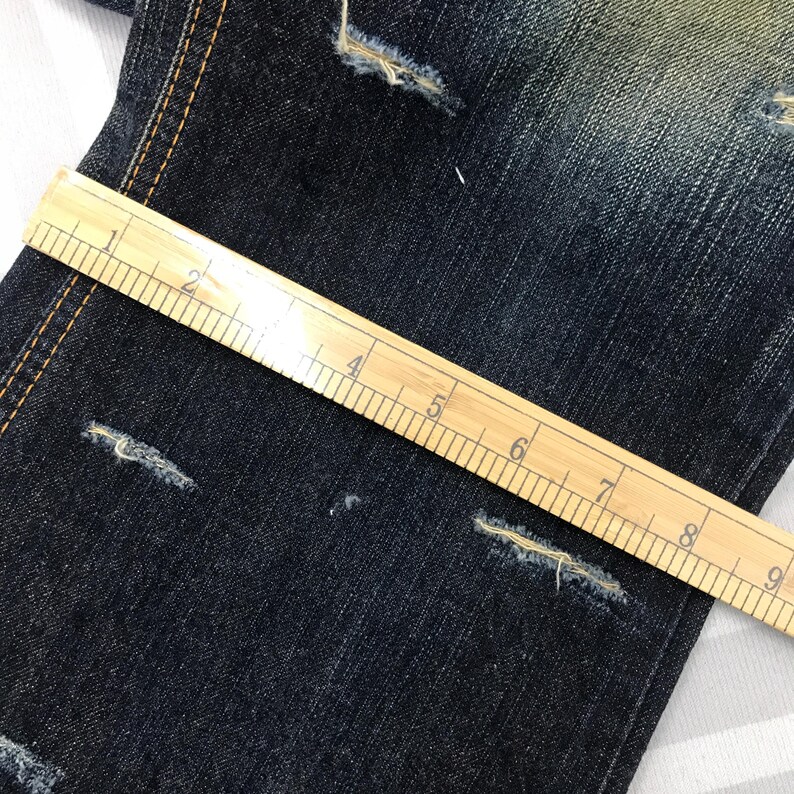 May include: Close-up of dark blue denim jeans with distressed details. A wooden ruler, marked in inches, is laid across the fabric, showing measurements. The jeans have visible wear and tear, with frayed edges.