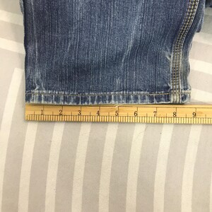 May include: Close-up of blue denim jeans with a wooden ruler measuring the hem. The ruler shows measurements in inches. The jeans have a classic five-pocket design and are made of durable denim fabric.