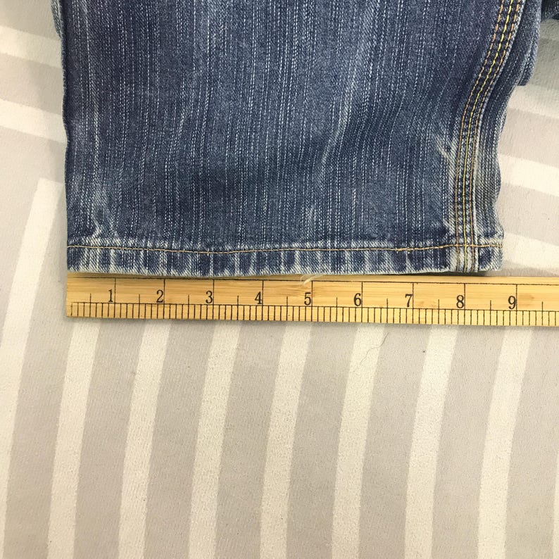 May include: Close-up of blue denim jeans with a wooden ruler measuring the hem. The ruler shows measurements in inches. The jeans have a classic five-pocket design and are made of durable denim fabric.