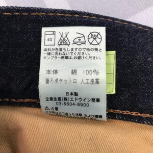 May include: Close-up of a denim garment label with Japanese text and care symbols. The label indicates the fabric is 100% cotton and provides washing instructions. The label also includes the manufacturer's information.