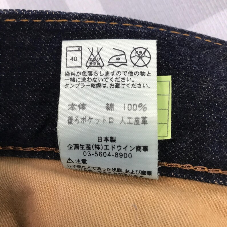 May include: Close-up of a denim garment label with Japanese text and care symbols. The label indicates the fabric is 100% cotton and provides washing instructions. The label also includes the manufacturer's information.