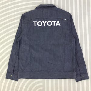 May include: A denim jacket in a dark blue hue, featuring the white word "TOYOTA" across the back. The jacket has a classic button-down design with two chest pockets and buttoned cuffs. The jacket is a size medium.