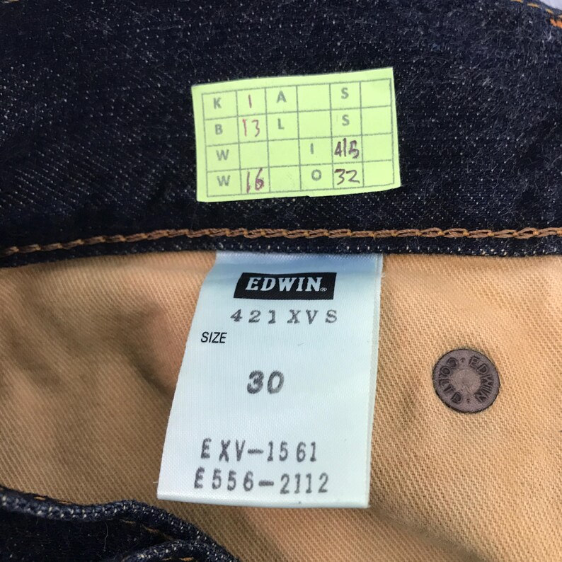 May include: Close-up of dark blue denim jeans with a visible label. The label reads "EDWIN" and "421 XVS", with a size of 30. Additional tags and a button are also visible.