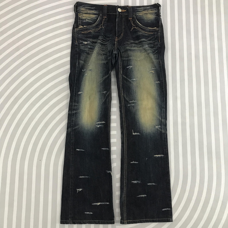 May include: Dark wash denim jeans with distressed detailing. The jeans feature a faded, worn look with intentional rips and light-colored patches. The design includes classic five-pocket styling and a button closure.