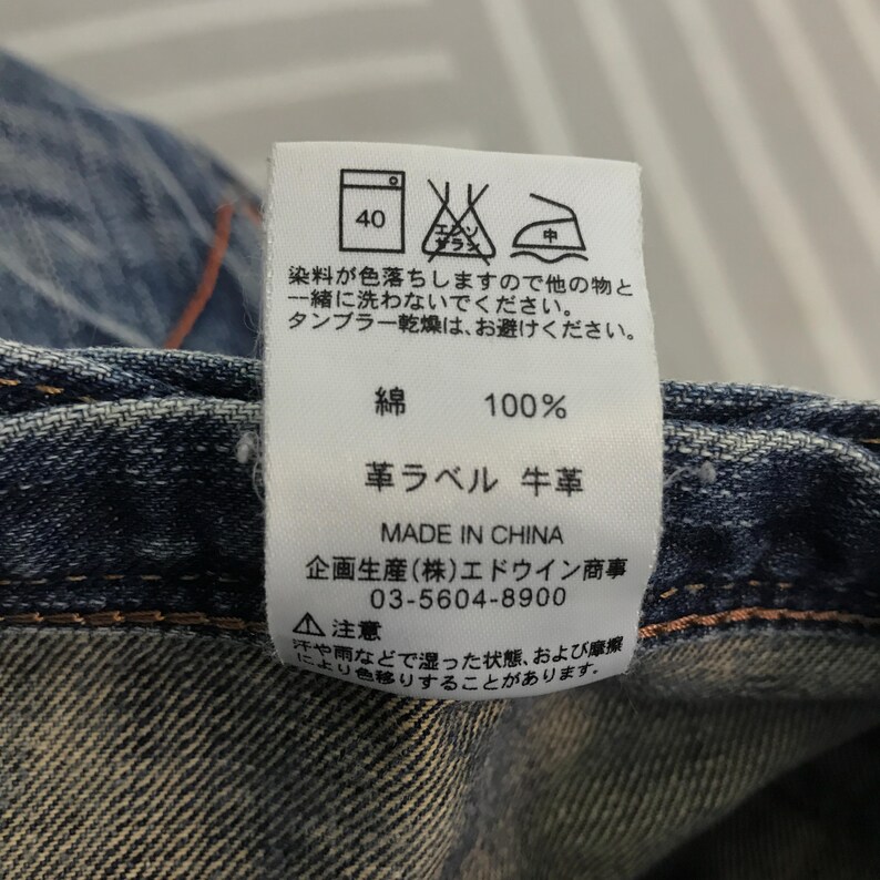 May include: Close-up of a denim garment with a white tag. The tag displays washing instructions, fabric composition (100% cotton), and the text "MADE IN CHINA". Additional text includes a phone number and warnings.