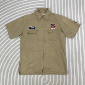 May include: A tan short-sleeved work shirt with a zip closure. The shirt has two patch pockets on the front and two embroidered patches. One patch says "Rebel" and the other has a red and white flag with a black X.