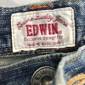 May include: Close-up of a blue denim garment featuring a white label with the text "Genuine Quality Jeans" and the brand name "EDWIN" in red. The label also includes the text "Exclusive design for lasting good look."