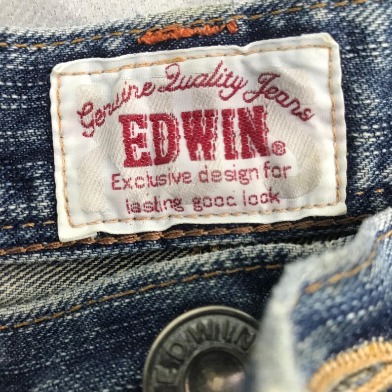 May include: Close-up of a blue denim garment featuring a white label with the text "Genuine Quality Jeans" and the brand name "EDWIN" in red. The label also includes the text "Exclusive design for lasting good look."