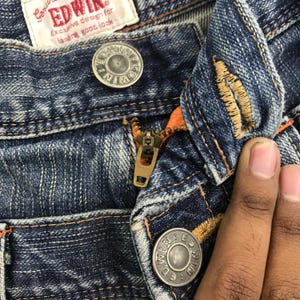 May include: Close-up of blue denim jeans with a zipper and button closure. The jeans have orange stitching and a metal zipper. The brand name "EDWIN" is visible on a tag.