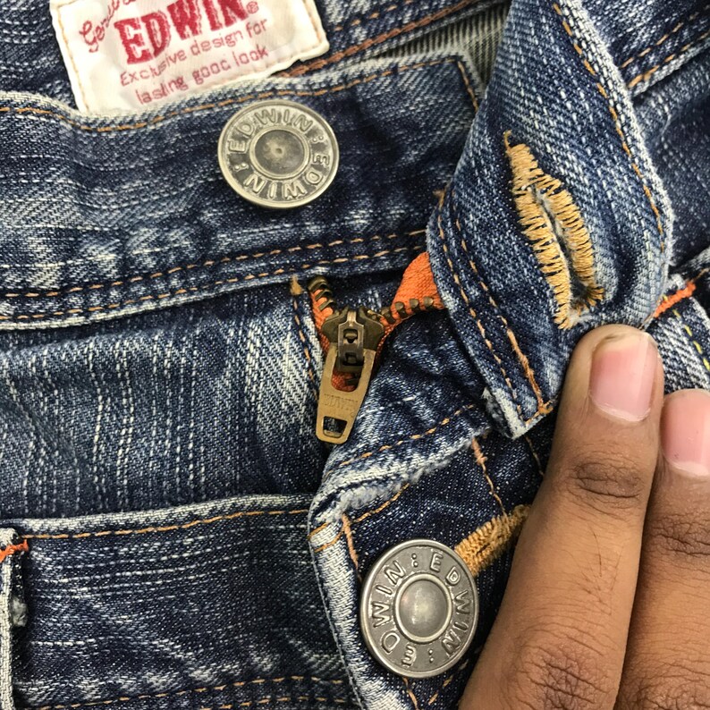 May include: Close-up of blue denim jeans with a zipper and button closure. The jeans have orange stitching and a metal zipper. The brand name "EDWIN" is visible on a tag.