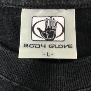 May include: Close-up of a black t-shirt tag with the "BODY GLOVE" logo in white. The logo features a hand inside an oval. The tag also has the size "-L-" printed in black.
