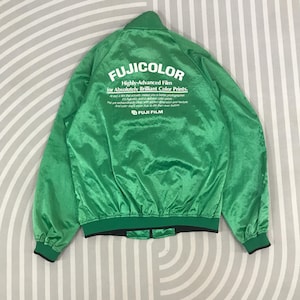 Puede incluir: Chaqueta bomber de satén verde con un logotipo blanco que dice "FUJICOLOR Highly-Advanced Film for Absolutely Brilliant Color Prints." El logotipo también incluye el texto "FUJI FILM" en una fuente más pequeña.