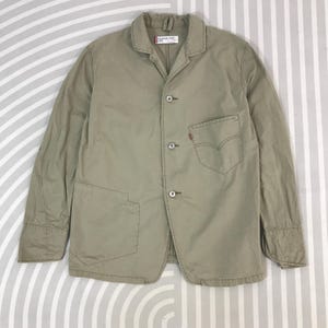 Puede incluir: Chaqueta beige con cuello, tres botones y dos bolsillos delanteros. La chaqueta tiene una pequeña etiqueta roja en el bolsillo delantero. Fabricada con un material ligero.