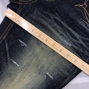 May include: Close-up of distressed blue denim jeans with a wooden ruler laid across the fabric. The ruler shows inch measurements from 1 to 10. The jeans have several small tears and faded areas.